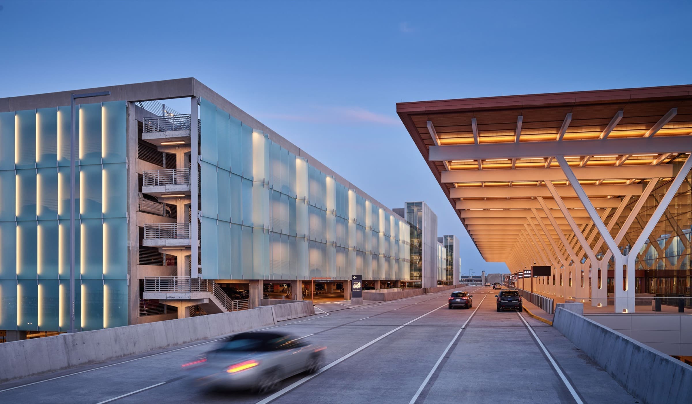 Kansas City International Airport at dusk, the parking structure clad in a backlit translucent glass rainscreen beside the terminal canopy